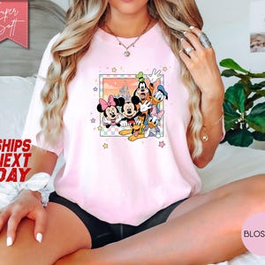 Może przedstawiać: Jasnor&oacute;żowa koszulka z kolorową grafiką postaci Disneya: Myszki Miki, Myszki Minnie, Goofy'ego, Kaczora Donalda, Daisy i Plutona. Koszulka ma napis "Super Soft" i "Ships Next Day".
