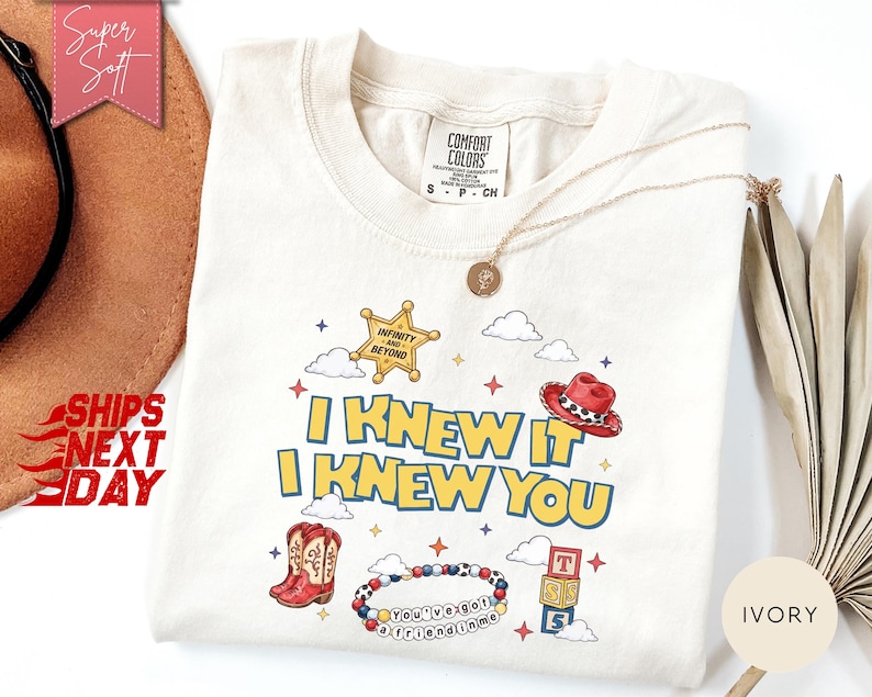 May include: An ivory t-shirt featuring a graphic print with the text I KNEW IT I KNEW YOU, a yellow sheriff star with the text INFINITY AND BEYOND, a red cowboy hat, red cowboy boots, a beaded bracelet with the text YOU'VE GOT A FRIEND IN ME, and blocks.