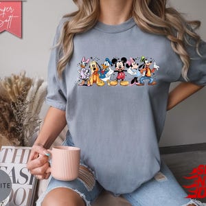 Puede incluir: Camiseta azul claro con un estampado colorido de personajes de Disney, incluyendo Mickey y Minnie Mouse. La camiseta tiene el texto "Super Soft" en una pancarta rosa. Una prenda c&oacute;moda.