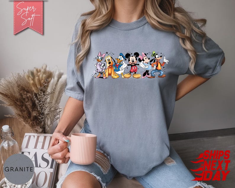 Puede incluir: Camiseta azul claro con un estampado colorido de personajes de Disney, incluyendo Mickey y Minnie Mouse. La camiseta tiene el texto "Super Soft" en una pancarta rosa. Una prenda c&oacute;moda.