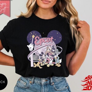 May include: Black t-shirt featuring a colorful graphic of Disney characters and the word "Epcot" in a stylized font. The design includes Mickey Mouse, Donald Duck, and other characters. The shirt has a round neckline and short sleeves.