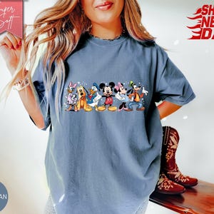 Puede incluir: Camiseta de color azul vaquero con un gr&aacute;fico colorido de personajes de Disney. El gr&aacute;fico incluye Mickey y Minnie Mouse, el Pato Donald, Goofy, Daisy Duck y Pluto. La camiseta tiene las palabras "Super Soft" y "Ships Next Day".