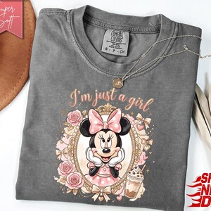 Może przedstawiać: Ciemnoszary t-shirt z kr&oacute;tkim rękawem z grafiką Myszki Minnie w ozdobnej złotej ramce, otoczonej r&oacute;żowymi r&oacute;żami i kokardkami. Nad ramką znajduje się napis "I'm just a girl" oraz mała ilustracja mrożonej kawy.