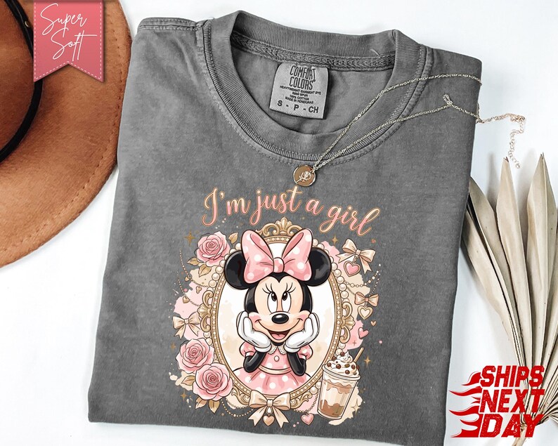 Może przedstawiać: Ciemnoszary t-shirt z kr&oacute;tkim rękawem z grafiką Myszki Minnie w ozdobnej złotej ramce, otoczonej r&oacute;żowymi r&oacute;żami i kokardkami. Nad ramką znajduje się napis "I'm just a girl" oraz mała ilustracja mrożonej kawy.