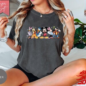 Puede incluir: Camiseta gris oscuro con un gr&aacute;fico colorido de personajes de Disney, incluyendo Mickey Mouse, Minnie Mouse, Donald Duck, Daisy Duck, Goofy y Pluto. La camiseta tiene cuello redondo y mangas cortas. El texto "Super Soft" est&aacute; en una pancarta rosa.