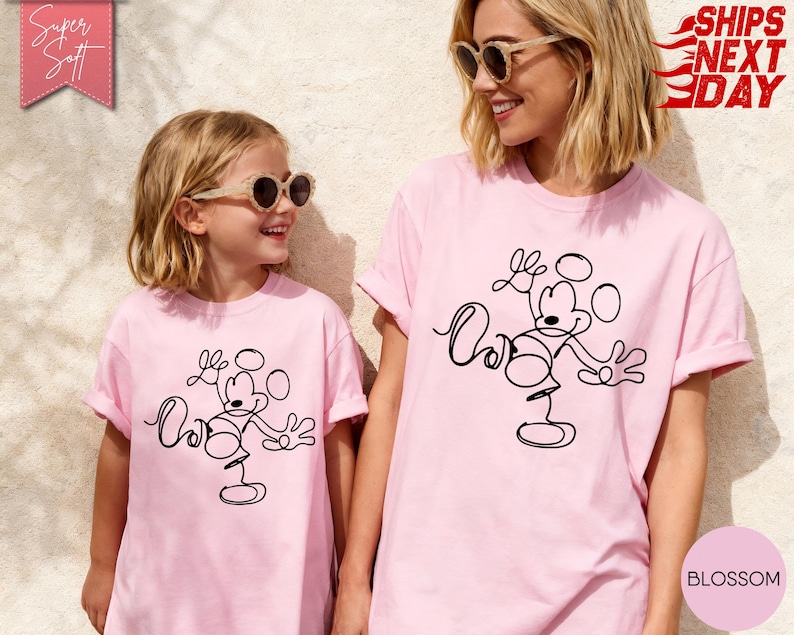 May include: Two people wearing matching light pink t-shirts with a black line drawing of Mickey Mouse. The shirts have rolled-up sleeves. The text "Super Soft" is in a pink banner in the upper left corner. The text "Ships Next Day" is in a red banner in the upper right corner.