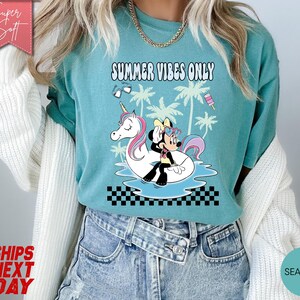 May include: A seafoam green t-shirt featuring a graphic of Minnie Mouse sitting on a white unicorn pool float in blue water. The design includes palm trees, sunglasses, a popsicle, and the text SUMMER VIBES ONLY above a black and white checkerboard pattern.
