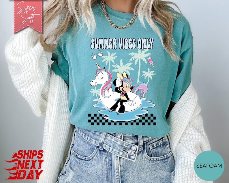 May include: A seafoam green t-shirt featuring a graphic of Minnie Mouse sitting on a white unicorn pool float in blue water. The design includes palm trees, sunglasses, a popsicle, and the text SUMMER VIBES ONLY above a black and white checkerboard pattern.