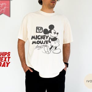 May include: Ivory-colored t-shirt featuring a black and white graphic of Mickey Mouse with the text "Mickey Mouse". The shirt has a classic, vintage design. The words "Super Soft" and "Ships Next Day" are also visible.