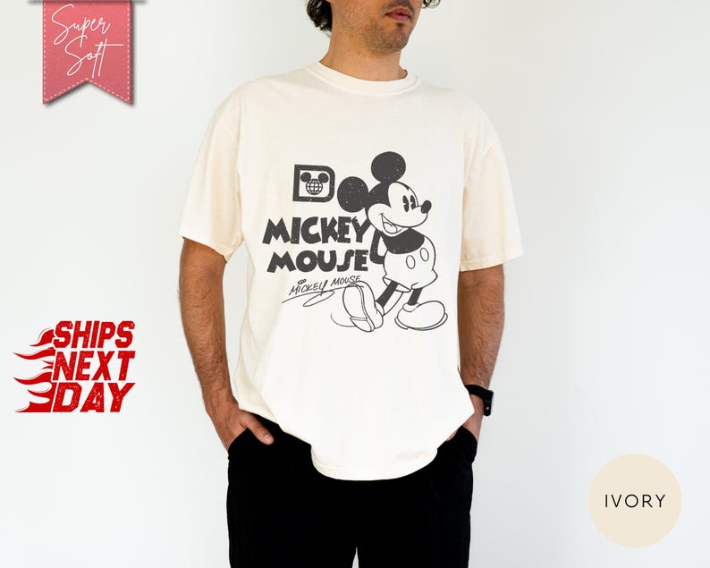 May include: Ivory-colored t-shirt featuring a black and white graphic of Mickey Mouse with the text "Mickey Mouse". The shirt has a classic, vintage design. The words "Super Soft" and "Ships Next Day" are also visible.