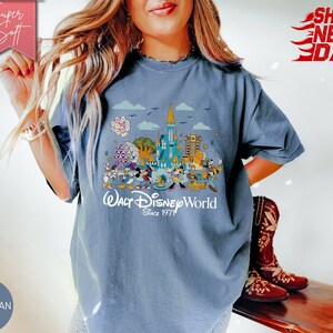 K&ouml;nnte beinhalten: Blaues Jeans-T-Shirt mit einer farbenfrohen Grafik von Walt Disney World-Charakteren und -Wahrzeichen. Das Design enth&auml;lt den Text "Walt Disney World Since 1971". Das Shirt hat kurze &Auml;rmel und einen Rundhalsausschnitt.