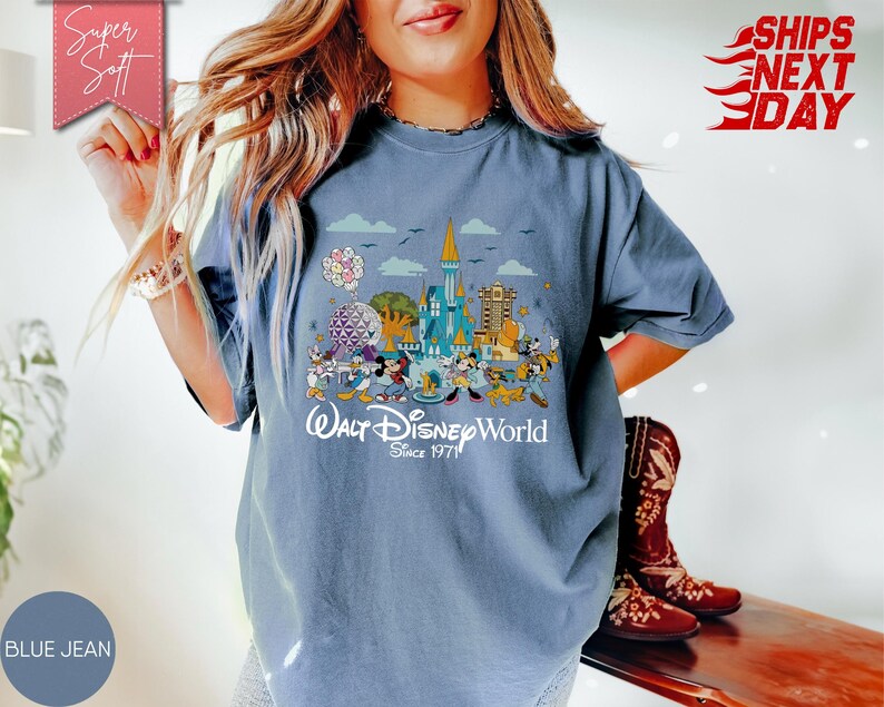 K&ouml;nnte beinhalten: Blaues Jeans-T-Shirt mit einer farbenfrohen Grafik von Walt Disney World-Charakteren und -Wahrzeichen. Das Design enth&auml;lt den Text "Walt Disney World Since 1971". Das Shirt hat kurze &Auml;rmel und einen Rundhalsausschnitt.