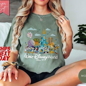 K&ouml;nnte beinhalten: Olivgr&uuml;nes T-Shirt mit einer farbenfrohen Grafik von Walt Disney World-Charakteren und -Wahrzeichen. Das Design umfasst Micky Maus, Donald Duck und das Schloss von Cinderella. Der Text "Walt Disney World Since 1971" ist darunter gedruckt.
