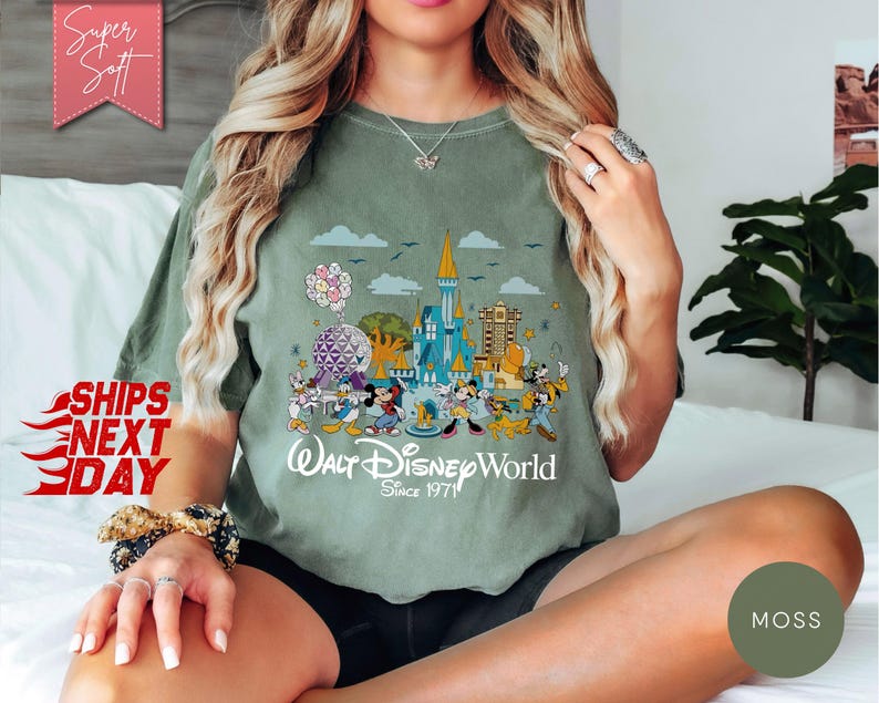 K&ouml;nnte beinhalten: Olivgr&uuml;nes T-Shirt mit einer farbenfrohen Grafik von Walt Disney World-Charakteren und -Wahrzeichen. Das Design umfasst Micky Maus, Donald Duck und das Schloss von Cinderella. Der Text "Walt Disney World Since 1971" ist darunter gedruckt.