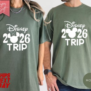May include: Two olive green t-shirts with the text "Disney 2026 TRIP" in white. One shirt features a Minnie Mouse design. The shirts have short sleeves and a crew neck. The words "Super Soft" and "Ships Next Day" are also visible.