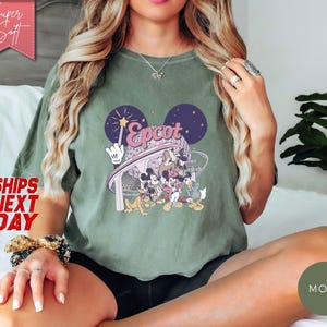 May include: Olive green t-shirt featuring a graphic of Mickey Mouse and friends at Epcot. The design includes the word "Epcot" in a stylized font, with a backdrop of stars and the iconic Mickey Mouse ears. The shirt has a relaxed fit.