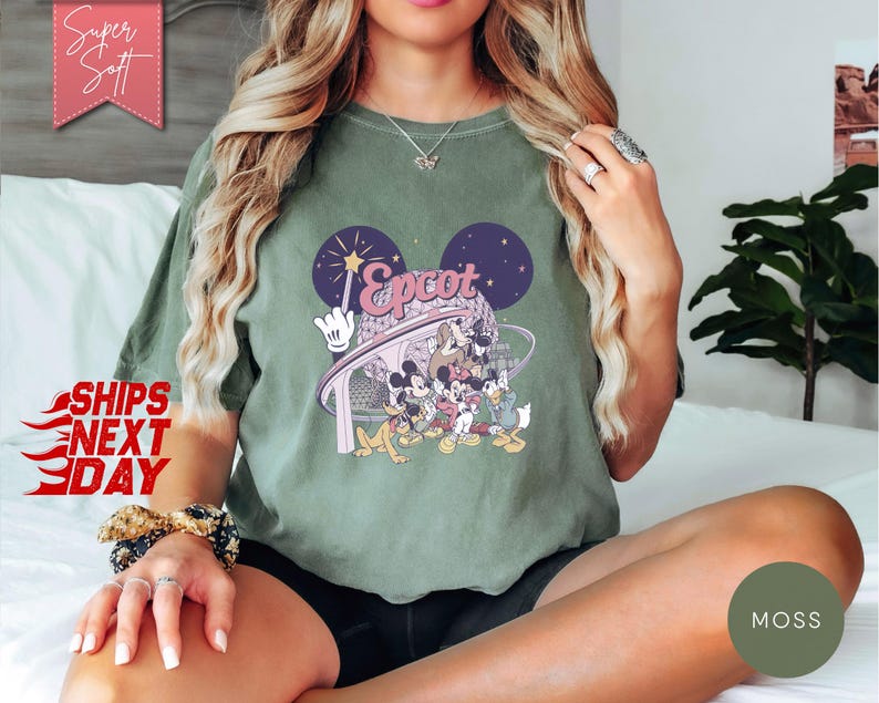 May include: Olive green t-shirt featuring a graphic of Mickey Mouse and friends at Epcot. The design includes the word "Epcot" in a stylized font, with a backdrop of stars and the iconic Mickey Mouse ears. The shirt has a relaxed fit.