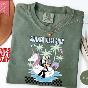 May include: A moss green t-shirt featuring a graphic of a cartoon mouse sitting on a white unicorn pool float in blue water. The design includes palm trees, sunglasses, a popsicle, and the text "SUMMER VIBES ONLY" in a light blue font.