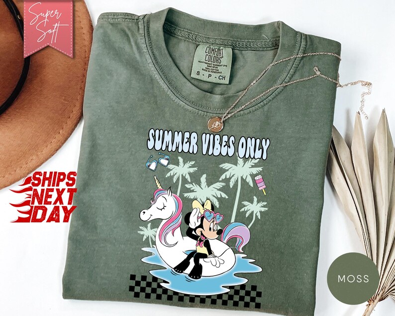 May include: A moss green t-shirt featuring a graphic of a cartoon mouse sitting on a white unicorn pool float in blue water. The design includes palm trees, sunglasses, a popsicle, and the text "SUMMER VIBES ONLY" in a light blue font.