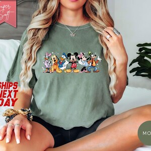 Puede incluir: Camiseta verde oliva con un gr&aacute;fico colorido de personajes de Disney, incluyendo Mickey y Minnie Mouse, Donald y Daisy Duck, Goofy y Pluto. La camiseta tiene el texto "Super Soft" y "Ships Next Day".