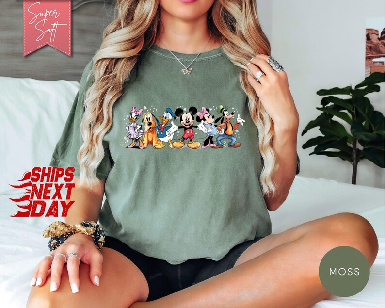Puede incluir: Camiseta verde oliva con un gr&aacute;fico colorido de personajes de Disney, incluyendo Mickey y Minnie Mouse, Donald y Daisy Duck, Goofy y Pluto. La camiseta tiene el texto "Super Soft" y "Ships Next Day".
