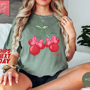 May include: Sage green t-shirt featuring a cartoon cherry design with Minnie Mouse bows and the Disney logo. The shirt has a relaxed fit and is made of soft material. The text "Super Soft" is in a pink banner.