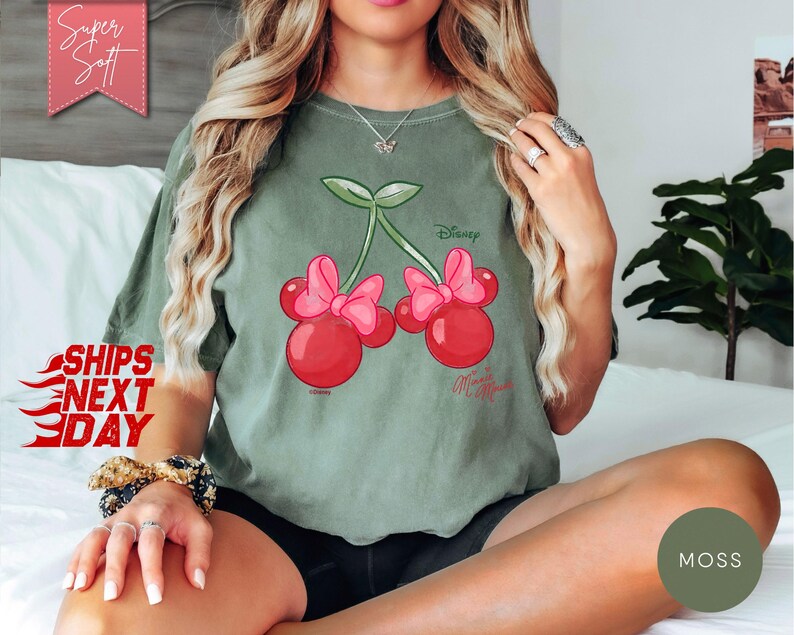 May include: Sage green t-shirt featuring a cartoon cherry design with Minnie Mouse bows and the Disney logo. The shirt has a relaxed fit and is made of soft material. The text "Super Soft" is in a pink banner.