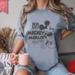 May include: A light blue t-shirt featuring a graphic of Mickey Mouse with the text "Mickey Mouse". The shirt has a vintage, distressed look. The words "Super Soft" are in a pink banner in the upper left corner.