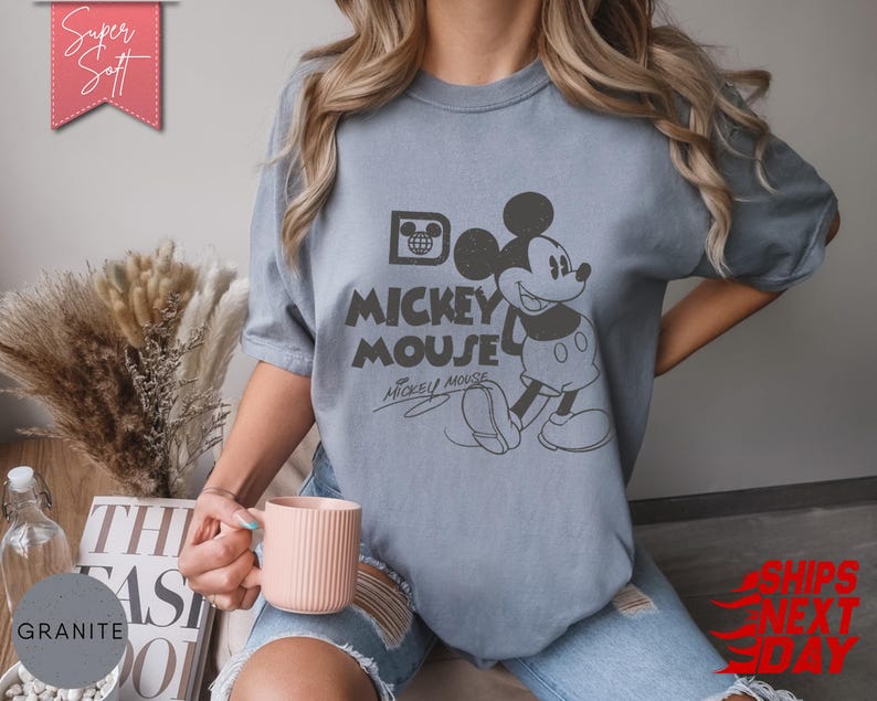 May include: A light blue t-shirt featuring a graphic of Mickey Mouse with the text "Mickey Mouse". The shirt has a vintage, distressed look. The words "Super Soft" are in a pink banner in the upper left corner.