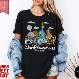 K&ouml;nnte beinhalten: Schwarzes T-Shirt mit einer farbenfrohen Grafik des Walt Disney World Schlosses, Figuren und der EPCOT-Kugel. Der Text "Walt Disney World Since 1971" ist darunter gedruckt. Das T-Shirt hat kurze &Auml;rmel.