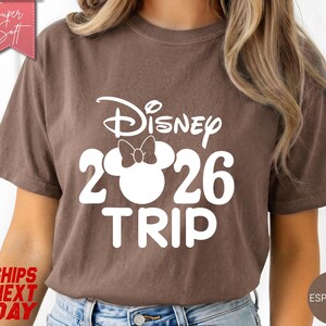 May include: Espresso-colored t-shirt with the text "Disney 2026 TRIP" in white. The design includes the Disney logo and a Mickey Mouse head with a bow. The shirt is a casual style.