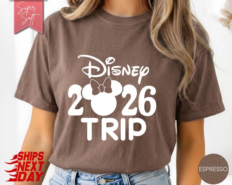 May include: Espresso-colored t-shirt with the text "Disney 2026 TRIP" in white. The design includes the Disney logo and a Mickey Mouse head with a bow. The shirt is a casual style.