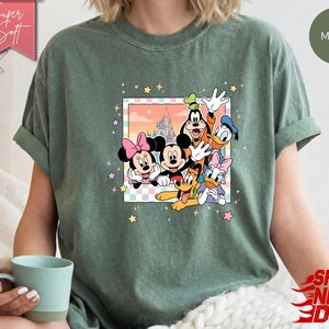 Może przedstawiać: Zielona koszulka w kolorze mchu z kolorową grafiką Myszki Miki, Myszki Minnie, Goofy'ego, Kaczora Donalda, Daisy Duck i Pluto. Grafika jest obramowana pastelową obw&oacute;dką i ilustracją zamku. Koszulka ma napis "Super Soft".