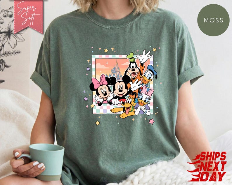 Może przedstawiać: Zielona koszulka w kolorze mchu z kolorową grafiką Myszki Miki, Myszki Minnie, Goofy'ego, Kaczora Donalda, Daisy Duck i Pluto. Grafika jest obramowana pastelową obw&oacute;dką i ilustracją zamku. Koszulka ma napis "Super Soft".