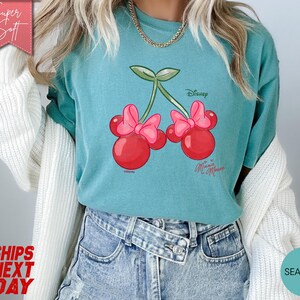 May include: Seafoam green t-shirt featuring a Minnie Mouse cherry design. The graphic shows two red cherries with pink bows and green stems. The shirt has the Disney and Minnie Mouse logos. The words "Super Soft" are in a pink banner.