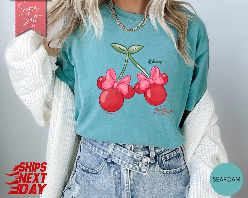 May include: Seafoam green t-shirt featuring a Minnie Mouse cherry design. The graphic shows two red cherries with pink bows and green stems. The shirt has the Disney and Minnie Mouse logos. The words "Super Soft" are in a pink banner.