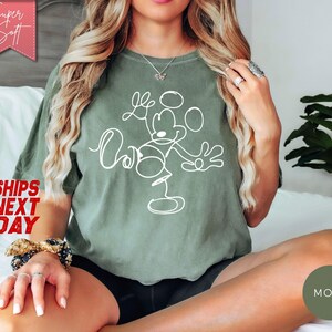 May include: Moss green t-shirt with a white line drawing of Mickey Mouse. The shirt has the words "Love" and "Oh" in a script font. The words "Super Soft" are in a pink banner in the upper left corner.