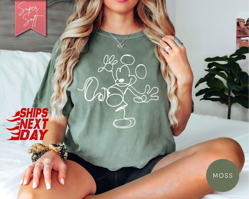 May include: Moss green t-shirt with a white line drawing of Mickey Mouse. The shirt has the words "Love" and "Oh" in a script font. The words "Super Soft" are in a pink banner in the upper left corner.
