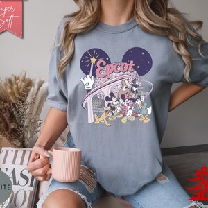 May include: Gray t-shirt featuring a colorful graphic of Disney characters, including Mickey Mouse, in front of a stylized Epcot design. The shirt has the text "Epcot" in pink and white. The shirt is made of a soft material.