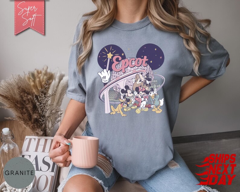May include: Gray t-shirt featuring a colorful graphic of Disney characters, including Mickey Mouse, in front of a stylized Epcot design. The shirt has the text "Epcot" in pink and white. The shirt is made of a soft material.