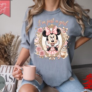 Może przedstawiać: Łupkowoniebieski t-shirt z grafiką Myszki Minnie w ozdobnej złotej ramce, otoczonej r&oacute;żowymi r&oacute;żami i kokardkami. Nad ramką znajduje się napis "I'm just a girl" wykonany kursywą oraz ilustracja mrożonej kawy.