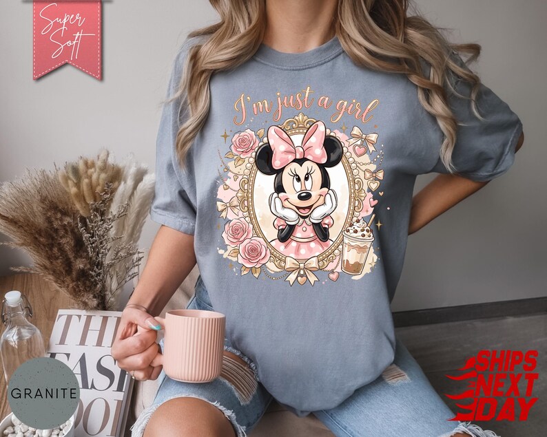 Może przedstawiać: Łupkowoniebieski t-shirt z grafiką Myszki Minnie w ozdobnej złotej ramce, otoczonej r&oacute;żowymi r&oacute;żami i kokardkami. Nad ramką znajduje się napis "I'm just a girl" wykonany kursywą oraz ilustracja mrożonej kawy.