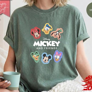 May include: Moss green t-shirt featuring Mickey Mouse and Friends. The shirt has colorful cartoon character patches of Mickey, Minnie, Donald, Daisy, and Goofy. The text "Disney MICKEY AND FRIENDS" is printed below the characters.