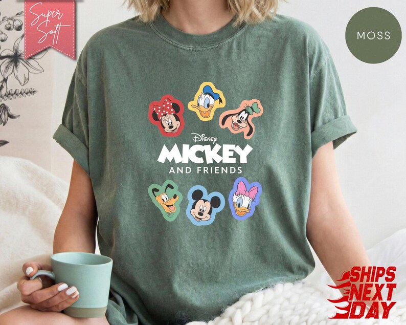 May include: Moss green t-shirt featuring Mickey Mouse and Friends. The shirt has colorful cartoon character patches of Mickey, Minnie, Donald, Daisy, and Goofy. The text "Disney MICKEY AND FRIENDS" is printed below the characters.