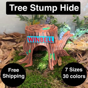 Puede incluir: Un escondite de tocón de árbol marrón con el texto "WINBELL" en un letrero azul. El escondite tiene un centro ahuecado y detalles verdes. La imagen también incluye el texto "Tree Stump Hide", "7 Sizes" y "30 colores".