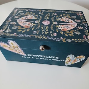 May include: A rectangular dark teal wooden box with a silver knob. The lid features a floral pattern with two moths. The front displays two heart-shaped floral motifs and the text: MONTPELLIER 90, 92 & 94 GRAND STREET. The box has a smooth finish.