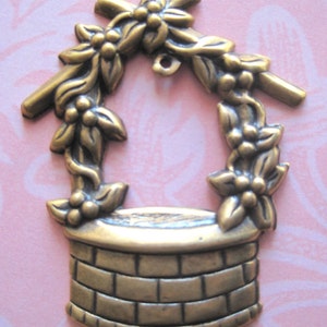 May include: A bronze-colored metal charm in the shape of a well. The well has a brick-like design and is topped with a floral arch. The charm has a small loop at the top for attaching to jewelry or other crafts.