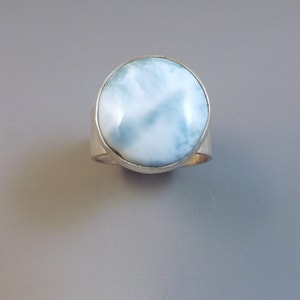 May include: A silver ring featuring a large, oval-shaped gemstone with a light blue and white marbled pattern. The gemstone is set in a silver bezel and has a polished appearance. The ring band is a simple, wide silver band.
