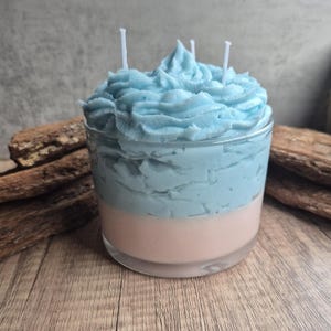 May include: A layered candle in a clear glass container. The bottom layer is a soft pink, topped with a light blue layer, and finished with a textured, whipped-cream-like light blue wax. Three white wicks are visible.