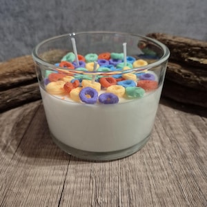 Puede incluir: Un portavelas de cristal transparente lleno de cera blanca y trozos de cereales de colores. Se ven dos mechas, listas para encender. La vela está sobre una superficie de madera, creando un artículo decorativo lúdico y único.
